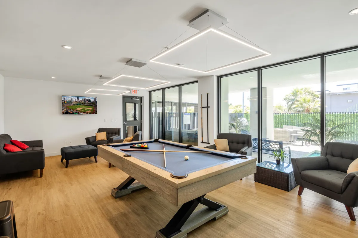 A stylish clubhouse interior featuring a pool table as the focal point, surrounded by comfortable sofas, a television, and large windows that let in natural light. The ambiance is casual and sociable, inviting residents to relax, play pool, and enjoy friendly gatherings.