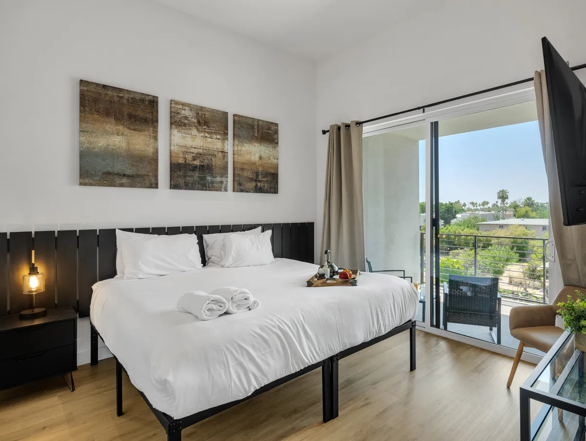 A king-size bed neatly made with soft white linens stands out at the center of a calming, softly lit bedroom. Decorative art pieces hang above the headboard, and a sliding glass door reveals a private balcony with outdoor seating and a tranquil view. Warm natural light fills the space, inviting relaxation and peaceful moments.