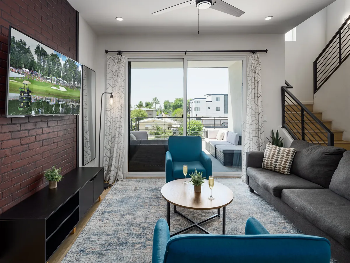 A contemporary living area with plush seating, a large flat-screen TV displaying a sports scene, and striking blue accent chairs. Floor-to-ceiling glass sliding doors open to a private balcony, blending indoor comfort with outdoor access.
