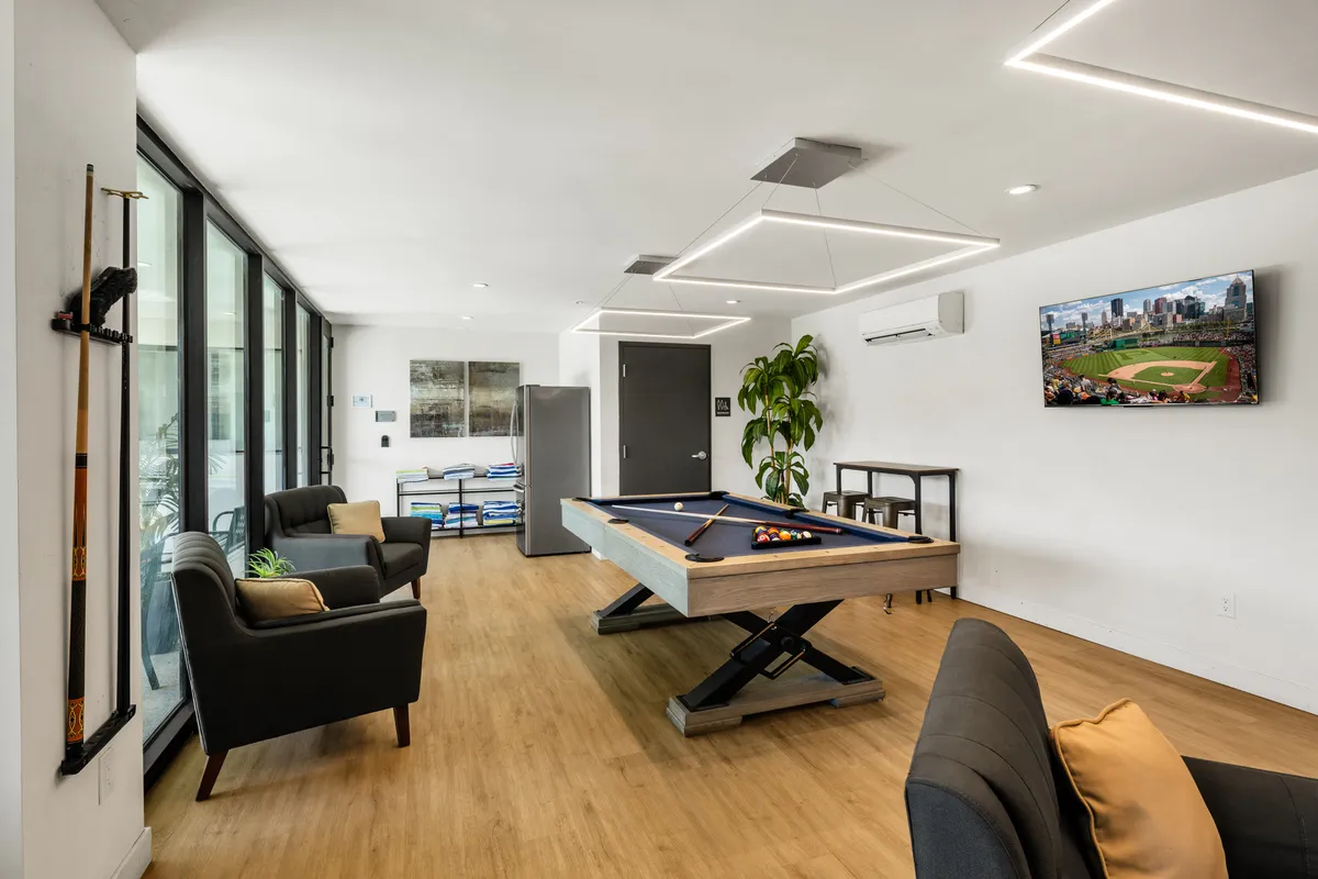 A modern community lounge featuring a pool table, plush sofas, a television, and greenery. The space invites social interaction and relaxation after a night out, perfectly tying the nightlife experience with upscale condo amenities.